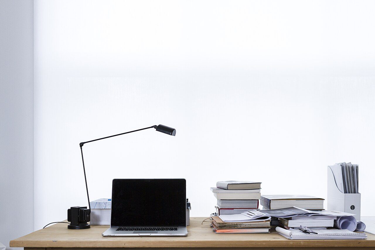 A laptop, books, paper files and lamp on a plain everyday desk